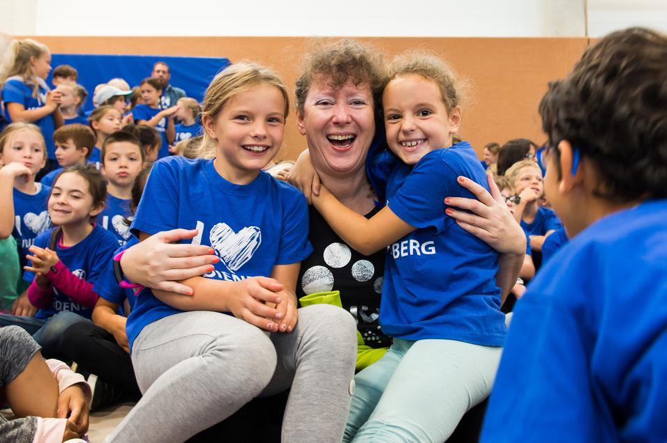 Lächelnde Kinder umarmen eine Frau in einem schwarzen Shirt, alle tragen blaue T-Shirts in einer Gruppenaktivität.