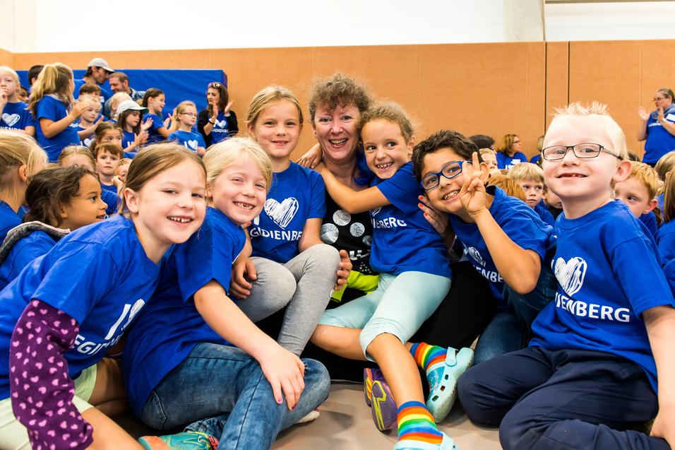 Gruppenkinder in blauen T-Shirts umarmen eine Frau, die lächelt, in einer Sporthalle voller Menschen.