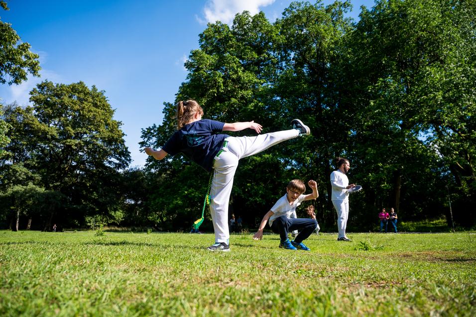 Zwei Kinder spielen im Park; eines tritt mit hohem Bein, das andere kniet und schaut überrascht. Grüne Bäume im Hintergrund.