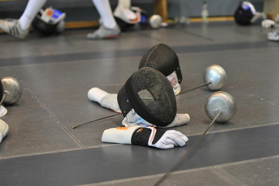 Zwei Fechthandschuhe und Masken liegen auf einer Matte, daneben stehen metallische Fechtwaffen.