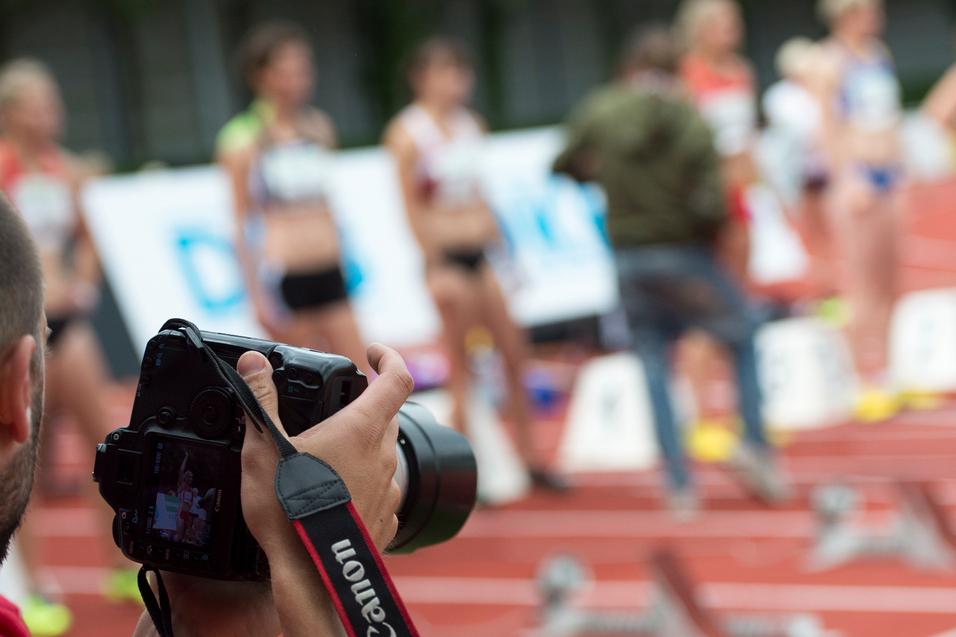 Fotograf mit Kamera im Vordergrund, im Hintergrund stehen Athletinnen an einer Laufbahn.