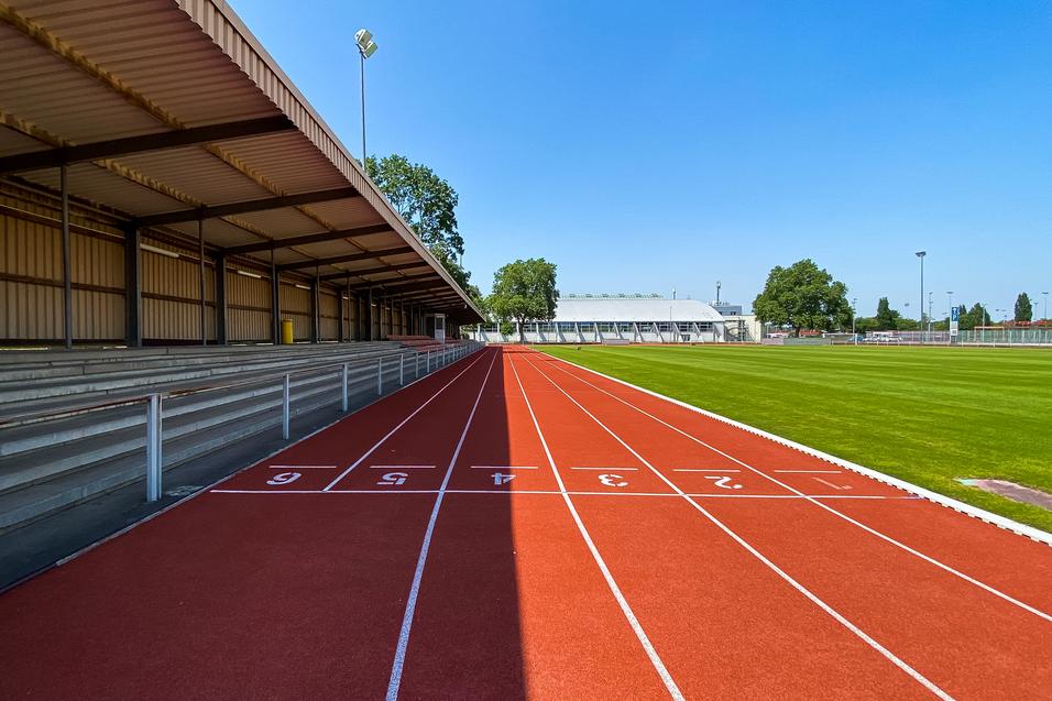 Leere Laufbahn mit roten Bahnen und Tribüne unter einem klaren blauen Himmel, neben einem Kunstrasenfeld.