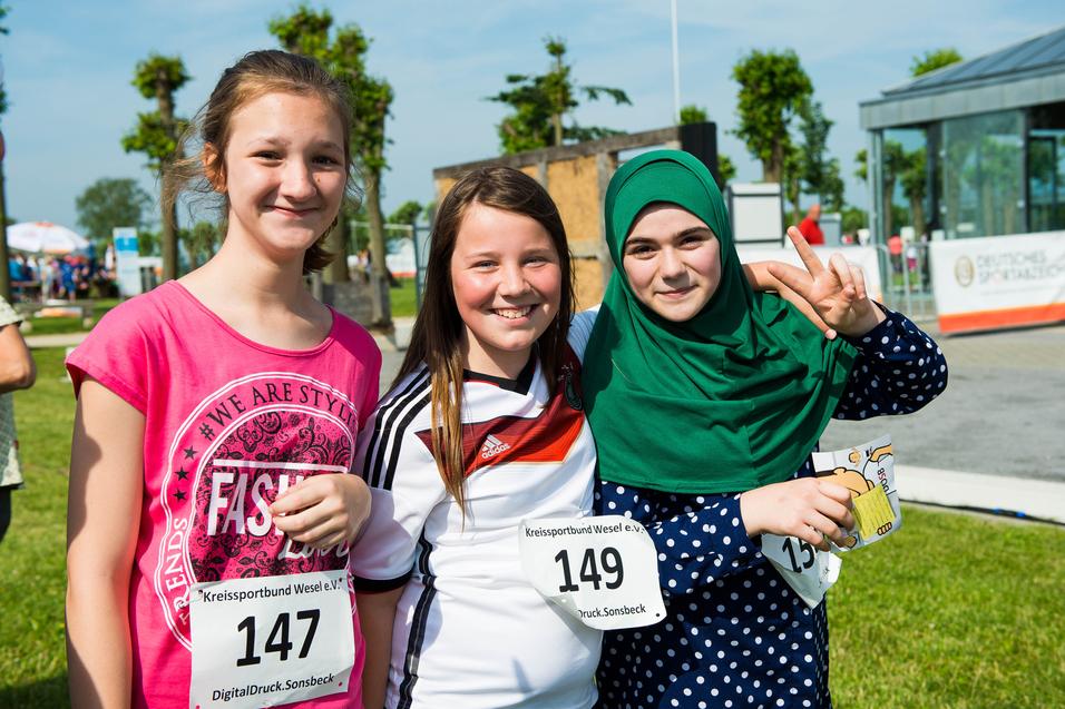 Drei lächelnde Mädchen in Sportkleidung mit Startnummern stehen auf einer Wiese, umgeben von einer Veranstaltung.