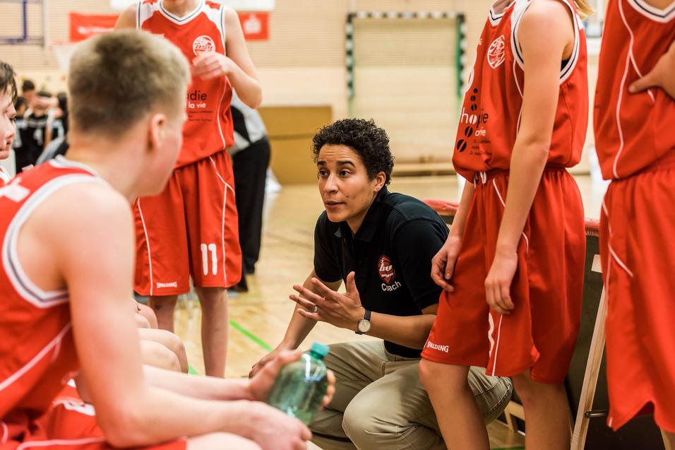Basketballtrainerin erklärt Taktik während einer Auszeit zu Jugendlichen in roten Trikots auf dem Spielfeld.