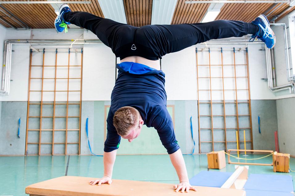 Junge macht Handstand auf einem Holztisch in einer Sporthalle mit verschiedenen Turngeräten im Hintergrund.