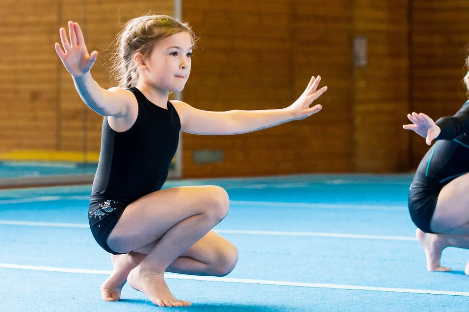 Zwei Mädchen in Gymnastikanzügen üben auf einer blauen Matte, eine kniet mit ausgestreckten Armen.