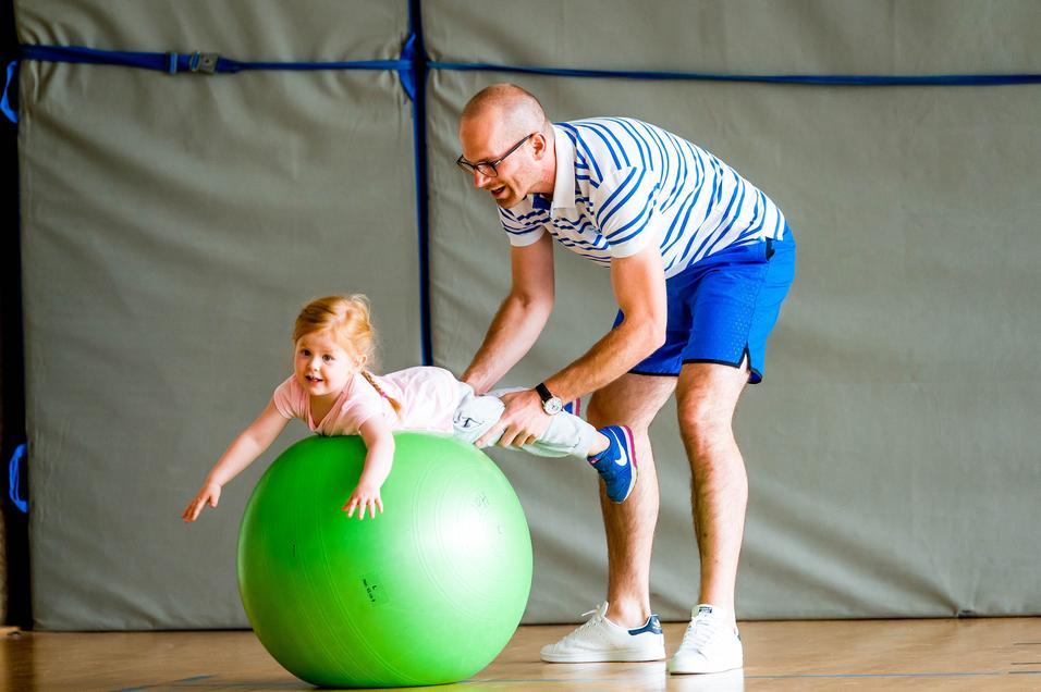 Ein Mann hält ein Kind, das auf einem grünen Gymnastikball sitzt, in einer Sporthalle. Beide lachen.