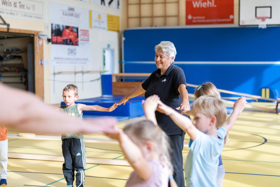 Seniorin leitet Kinder in einer Sporthalle an, während sie ihre Arme in die Höhe strecken.