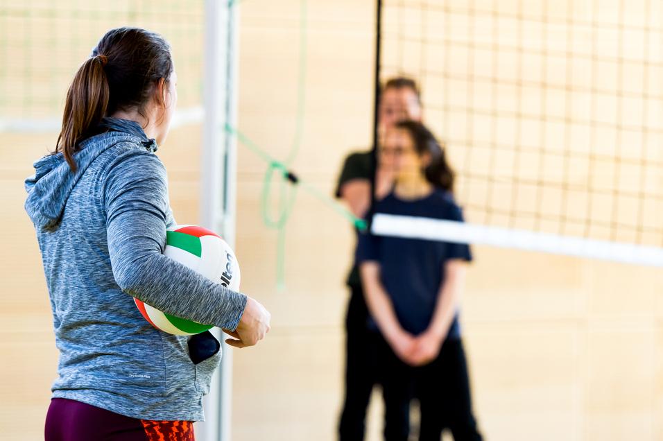 Eine Person mit einem Volleyball steht vor dem Netz, während zwei weitere Personen im Hintergrund warten.